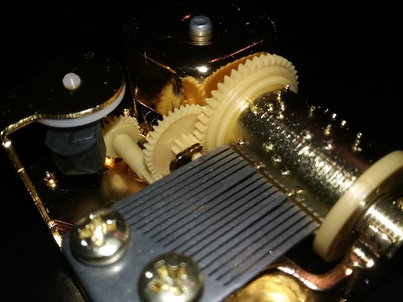 Op de afbeelding: Close-up van een goudkleurig muziekdoosmechanisme met een zwart-wit gestreepte kam en een reeks tandwielen. Het mechanisme is gemaakt van metaal en heeft een gepolijste afwerking.