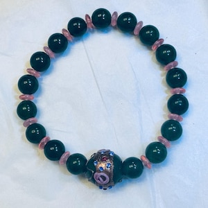 May include: A beaded bracelet featuring dark green, round beads interspersed with smaller, pink, oblong beads. A central, decorative bead is a darker green with a floral design and blue and pink accents. The bracelet is on a white background.