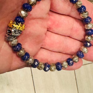 May include: A beaded bracelet featuring alternating blue and silver beads. The bracelet includes a silver skull charm wearing a gold crown. The bracelet is held in a hand.