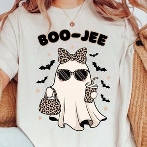 May include: Cream t-shirt with a cartoon ghost wearing sunglasses and a leopard print bow, holding a purse and a coffee cup with a straw. The text "BOO-JEE" is above the ghost, with bats and stars around it.