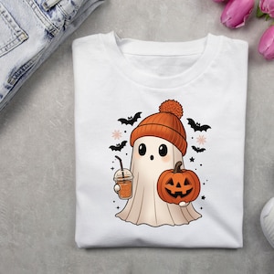 May include: White t-shirt featuring a cartoon ghost wearing an orange beanie and holding a pumpkin and a drink. Black bats and stars surround the ghost, creating a Halloween theme. The shirt is displayed with jeans and white sneakers.
