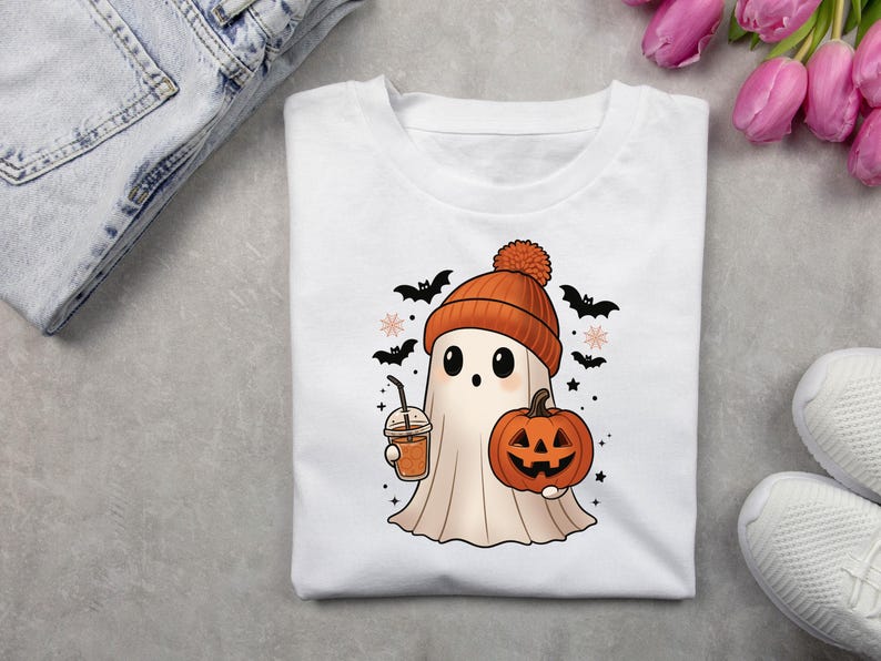 May include: White t-shirt featuring a cartoon ghost wearing an orange beanie and holding a pumpkin and a drink. Black bats and stars surround the ghost, creating a Halloween theme. The shirt is displayed with jeans and white sneakers.