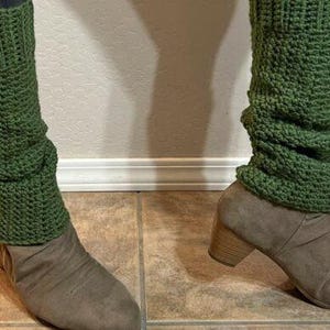 Puede incluir: Un par de calentadores de piernas de ganchillo verde usados sobre botines de ante grises. Los calentadores de piernas son de punto texturizado y las botas tienen un tacón bajo. El fondo es una pared neutra y un suelo embaldosado.