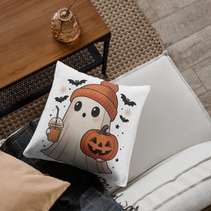 May include: White square pillow with a cartoon ghost wearing an orange beanie and holding a pumpkin and a drink. The pillow has a Halloween theme with bats and stars.