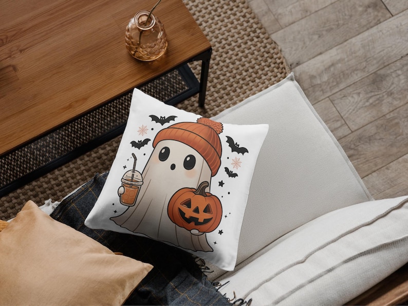 May include: White square pillow with a cartoon ghost wearing an orange beanie and holding a pumpkin and a drink. The pillow has a Halloween theme with bats and stars.