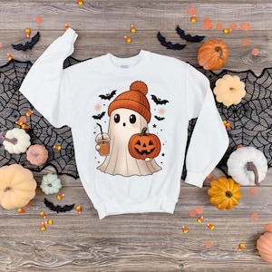 May include: White sweatshirt with a Halloween-themed graphic. The design features a cartoon ghost wearing an orange beanie, holding a pumpkin and a coffee drink. Black bats and pumpkins surround the design. The sweatshirt is surrounded by Halloween decorations.