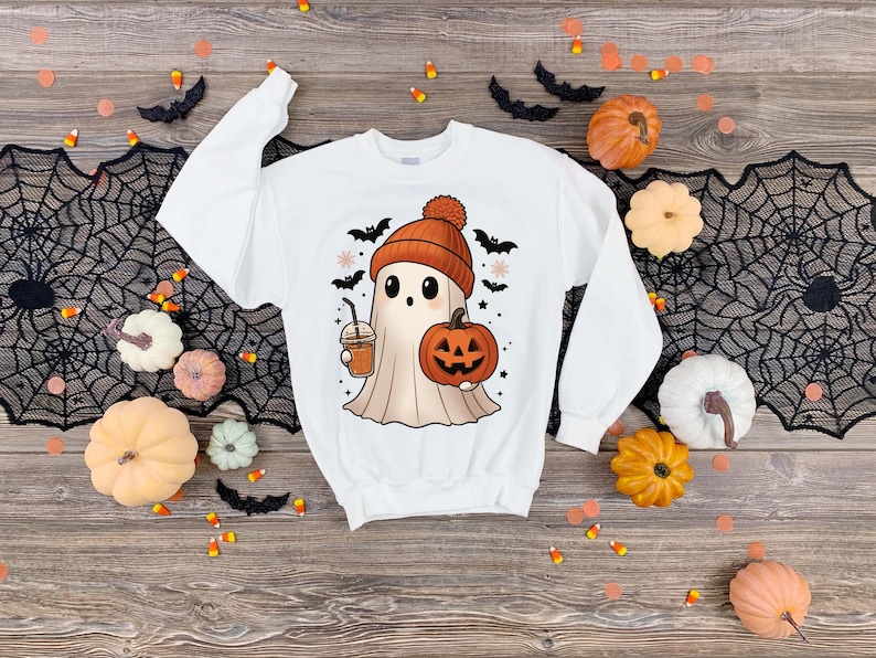 May include: White sweatshirt with a Halloween-themed graphic. The design features a cartoon ghost wearing an orange beanie, holding a pumpkin and a coffee drink. Black bats and pumpkins surround the design. The sweatshirt is surrounded by Halloween decorations.