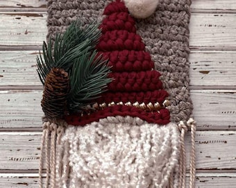 May include: A handmade Christmas gnome wall hanging. The gnome features a burgundy hat with a white pom-pom, a white beard, and a gray background. A pinecone and evergreen sprig are attached to the side. The wall hanging is made of yarn.