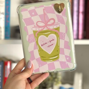 May include: Clear protective case for an e-reader with a green and pink design. The design features a matcha drink illustration with the text "Matcha + Books - Heaven" inside a heart. A gold heart-shaped charm is attached.