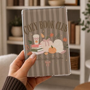 May include: A clear tablet case featuring a brown and beige vertical striped background with the text COZY BOOK CLUB. The design includes illustrations of a coffee cup, stacked books, pumpkins, and a yarn ball with small bows and autumn leaves.