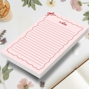 May include: A pink notepad with a scalloped edge and the word "notes" in a cursive font. The notepad has horizontal lines for writing and a small bow design at the top. The notepad is surrounded by flowers, leaves, and a glass of iced coffee.