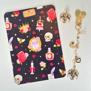 May include: A black decorative card with a repeating pattern of roses, skulls, books, and perfume bottles. A gold-colored dragon charm and a gold zipper pull with a heart and dangling charms are also present.