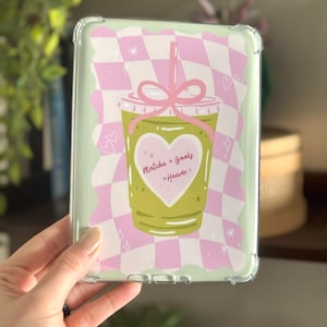 May include: Clear protective case for an e-reader with a graphic of a matcha drink in a to-go cup. The cup has a pink bow and the words "Matcha + Books = Heaven" in a heart. The background is a pink and white checkered pattern.