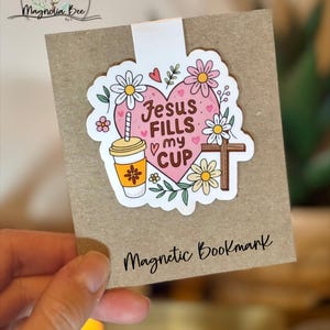 May include: A magnetic bookmark featuring a heart-shaped design with the text "Jesus Fills my Cup." The design includes a coffee cup, cross, and floral elements. The bookmark is attached to a brown card.