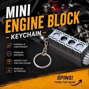 May include: A gray and black keychain shaped like a miniature four-cylinder engine block. The object features a silver-colored metal ring attached to one end and a small, light gray gear on the opposite side that rotates. The surface has a textured finish.
