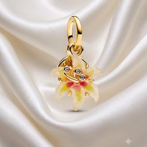 Pode incluir: Um pingente de tom dourado em forma de flor de lírio com pétalas cor de creme, um centro rosa e amarelo e três pequenas pedras transparentes incrustadas no meio. O pingente possui uma argola circular no topo para fixação em uma corrente.