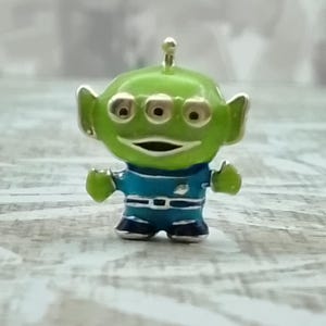 Peut inclure: Une petite breloque en métal représentant un personnage extraterrestre à trois yeux avec une peau vert vif, un uniforme bleu avec une ceinture argentée et une petite antenne sur le dessus de la tête, une bouche souriante et des oreilles pointues.