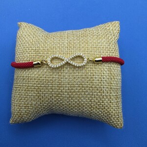 Peut inclure: Un bracelet en cordon rouge avec un symbole infini doré au centre, orné de petites pierres transparentes. Le bracelet est présenté sur un coussin en tissu beige texturé, mettant en valeur le contraste entre le cordon rouge vif et le métal doré.