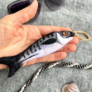 Puede incluir: Un llavero de pez de fieltro en tonos grises, negros y blancos, con un cierre de metal dorado. El pez tiene un diseño detallado, incluyendo un ojo y marcas. Gafas de sol y un collar de cuentas en el fondo.