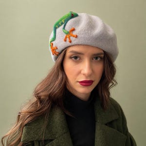 Może przedstawiać: Jasnoszary beret z zielono-pomarańczowym wzorem żaby. Żaba jest wyhaftowana z przodu kapelusza, z zielonym korpusem, pomarańczowymi stopami i czerwonymi oczami. Beret wykonany jest z miękkiego materiału przypominającego filc.