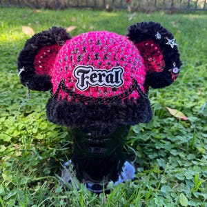May include: A pink and black crochet hat with ear-like extensions. The hat features the word "Feral" in a white and black patch, a black chain, and star-shaped charms. The hat is displayed on a black mannequin head.