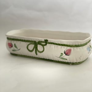 May include: Cream-colored rectangular ceramic planter with a green rope design around the rim. The planter is decorated with green bows and pink and blue floral accents. A decorative piece for indoor or outdoor use.