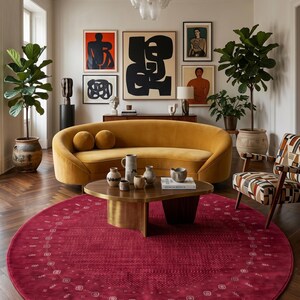May include: A modern living room with a mustard yellow sofa, a round coffee table, and a patterned armchair. A large, round, red rug sits on the hardwood floor. Artwork hangs on the wall above the sofa. The room is decorated with plants and a chandelier.