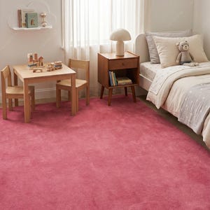 May include: A large, solid-colored, deep pink area rug with a soft, plush texture covering the floor of a bedroom. The rug features a uniform, dense pile that extends across the room, positioned beneath a wooden table, chairs, and a bed.