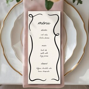 Peut inclure: Mise en place élégante avec des couverts dorés, des assiettes blanches à bord doré et une serviette rose. Une carte de menu avec du texte noir et des lignes décoratives énumère les plats.
