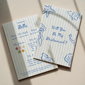 May include: Two rectangular cards with a light blue and white vertical striped background. The cards feature blue line art of a starfish, jellyfish, fish, anchor, and shell. Text includes "Will You Be My Bridesmaid?" and "bridesmaid Bella".
