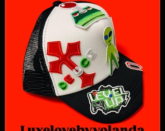 Op de afbeelding: Zwarte en witte truckerpet met een wit voorpaneel met rood en groen geborduurde patches. De patches bevatten een alien, een joystick en het woord "SPACE". De pet heeft een zwarte mesh achterkant en een gebogen klep.