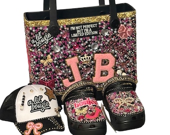 May include: A black tote bag, a pair of black clogs, and a white and black trucker hat, all decorated with pink, gold, and clear rhinestones, pearls, and decorative accents. The tote bag has the text "I'M NOT PERFECT BUT I'M A LIMITED EDITION". The clogs and hat have the text "a lil bougie".