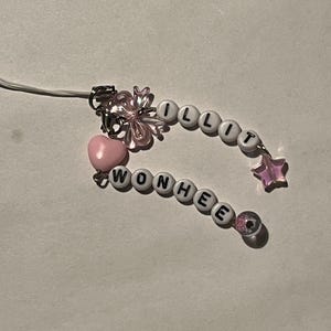 May include: A pink and white beaded charm with the names "ILLIT" and "WONHEE". The charm includes a pink heart, a clear flower, a pink star, and a clear bead. The charm is attached to a white cord.