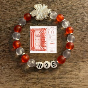 May include: A bracelet with alternating red and frosted glass beads, separated by silver-coloured spacers. The bracelet has white letter beads spelling "W2E" and a white bow-shaped charm. A small diagram is also included.
