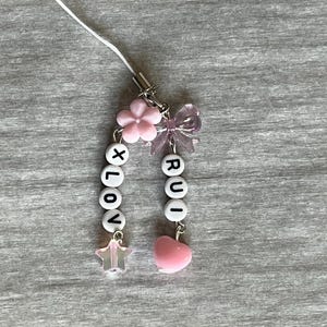 May include: A handmade charm with the words "XLOV" and "RUI" spelled out in white letter beads. The charm features pink flower and bow accents, a star, and a pink heart charm. The charm is attached to a white cord.