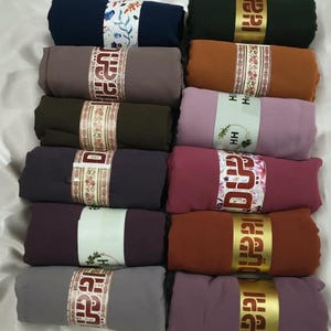 May include: A collection of ten rectangular fabric scarves arranged in two columns. Each scarf is folded into a compact roll and secured with a paper band featuring floral patterns and the text HH. The scarves are in various solid colours including navy, brown, olive, plum, grey, black, tan, mauve, maroon, and rust.