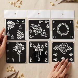 May include: A set of six black adhesive stencils for temporary body art, featuring white cut-out designs of roses, floral arrangements, a circular wreath, crescent moons, and hanging lantern motifs, organized in clear plastic sleeves on a light background.