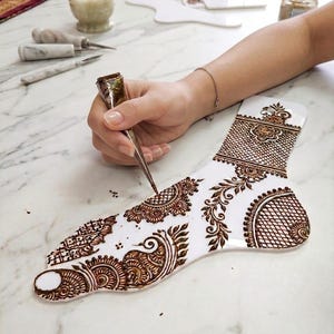 Puede incluir: Un esténcil blanco con forma de pie con intrincados diseños de henna marrón se está rellenando con un cono de henna. El arte del henna presenta patrones florales y geométricos. La imagen está tomada sobre una superficie de mármol.