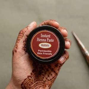 Puede incluir: Un pequeño recipiente negro y redondo de pasta de henna instantánea color granate. La etiqueta presenta el texto "Instant Henna Paste", "MAROON" y "Permissible Skin Friendly" en letras blancas y amarillas sobre un fondo rojo oscuro.