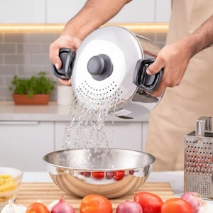 Puede incluir: Una olla de acero inoxidable con tapa colador se sostiene sobre un cuenco de acero inoxidable, con agua saliendo. La olla tiene asas negras y un pomo gris. Tomates rojos y cebollas moradas están sobre una tabla de cortar de madera.