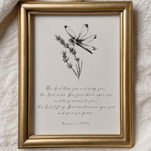 May include: A framed print with a gold-colored frame. The artwork features a black and white illustration of a dragonfly and lavender. The print includes a script with the text "The Lord bless you and keep you...Numbers 6:24-26."