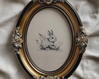 May include: An oval framed print featuring a detailed pen and ink drawing of a rabbit in a grassy setting. The frame is ornate, with a gold inner edge, black outer edge, and floral embellishments.