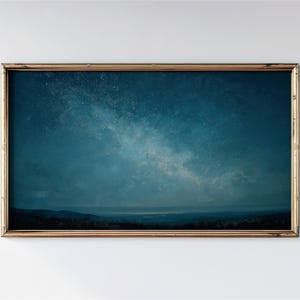 May include: A framed print featuring a celestial landscape. The artwork depicts a dark blue night sky filled with stars, with a lighter, milky way-like band across the center. The bottom of the image shows a dark silhouette of a landscape. The frame is gold.