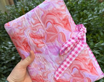 May include: A quilted notebook cover with a vibrant, swirled pattern in shades of pink and red. The cover is secured with a pink and white gingham ribbon tie. The notebook cover is held in a hand.
