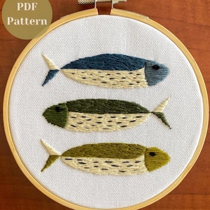 May include: A circular wooden embroidery hoop containing white fabric with a hand-stitched design of three fish stacked vertically. The fish are rendered in shades of dark blue, forest green, and olive green with cream-colored bellies and black details.