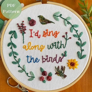 Può includere: Ricamo su telaio con la scritta "I'd sing along with the birds" nei colori dell'arcobaleno. Il disegno comprende uccelli ricamati, fiori e verde. Un'etichetta del modello PDF è visibile in alto a sinistra.