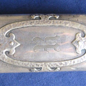 Könnte beinhalten: Ein silberfarbenes, rechteckiges Etui mit dekorativen kreuzförmigen Akzenten und einem ovalen Mitteldesign. Das Etui hat eine eingravierte Initiale und gehämmerten Metalldetails, was auf einen Vintage- oder antiken Stil hindeutet. Ca. 15 cm lang.