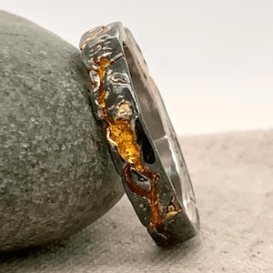 May include: A silver ring with a textured, dark gray exterior and gold-colored accents. The ring is leaning against a gray stone, showcasing its unique design and craftsmanship. The gold accents add a touch of elegance to the ring.