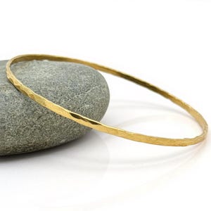May include: A gold-colored bangle bracelet with a hammered texture. The bracelet is circular and rests on a gray stone against a white background. The bangle is a simple, elegant piece of jewelry.