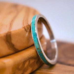 Puede incluir: Un anillo de plata con una incrustación de turquesa descansa sobre una superficie de madera. La banda del anillo es delgada y la turquesa tiene un aspecto moteado. La veta de la madera es visible, con tonos marrones cálidos.